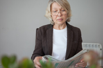 An elegant woman in glasses sits at a glass table and holds a newspaper in her hands.