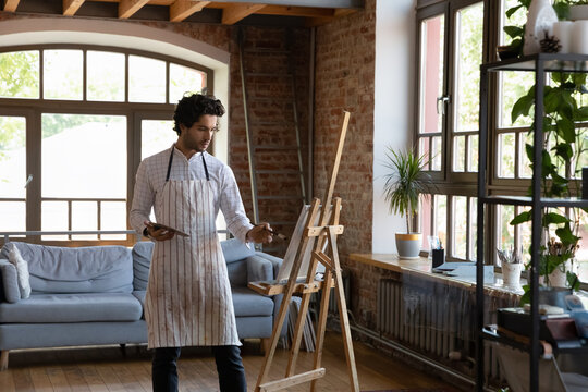 Serious millennial 30s focused man painter holding palette use paintbrush painting on canvas standing alone in loft workshop engaged in creative hobby. Professional occupation person, creation concept