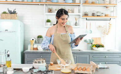 young Asian housewife in kitchen using tablet for searching recipes online cooking the bakery dough homemade.
