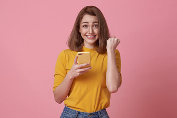 Happy young woman using mobile phone and hand gesture success celebrate isolated over pink...