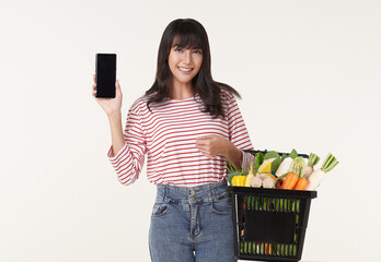 Happy Asian woman showing smartphone holding basket full of fresh vegetable groceries isolated on...
