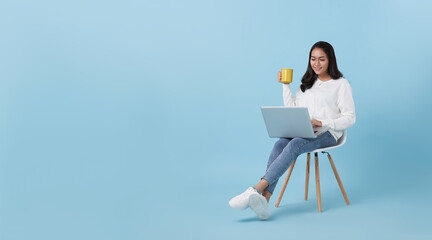 Young woman asian happy smiling in casual white cardigan with denim jeans.While her using computer laptop sitting on white chair and drinking coffee isolate on bright blue background. - Powered by Adobe