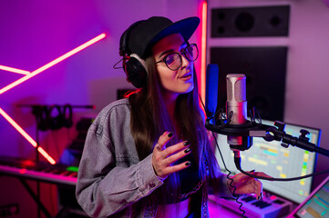 Beautiful stylish girl singing into a microphone in a recording studio with neon lights in a cap...