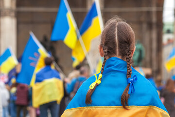 Rally support Ukraine in Europe. Ukraine Flag on Shoulders of Girl with blue yellow ribbons on flags background. Stand with Ukraine concept. 