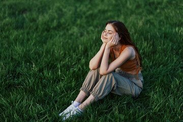 A beautiful girl relaxing in nature sitting in pants and top on the green grass in the rays of the setting summer sun. The concept of health and care for the body and the environment 