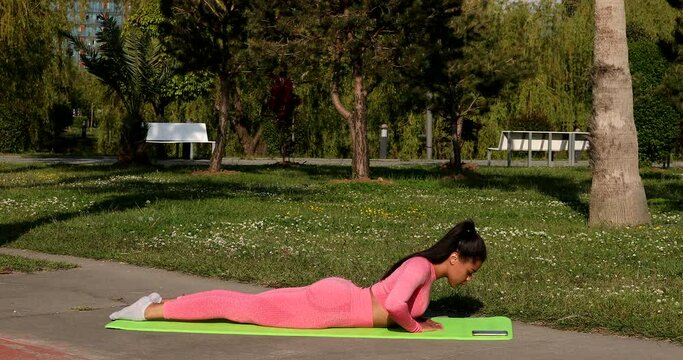Young woman doing yoga practice or stretching on green grass in park. Side view of black female does asana or posture and looks with smile, stretches legs, arms and poses for yoga.