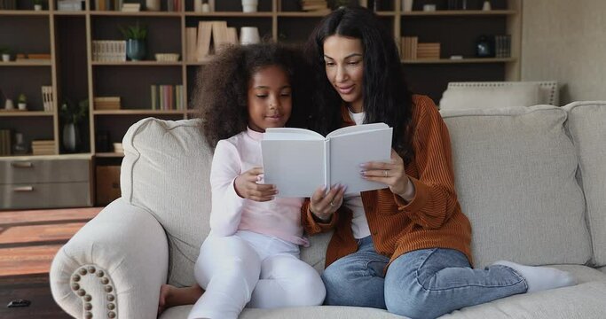 African mom and little daughter spend leisure read book, sit on sofa in cozy living room look through fairy tale pictures looking happy enjoy favourite hobby. Children growth and development concept