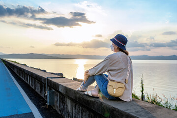 Female tourist looking away at river and mountain. Woman traveler sitting on concrete of lake or dam with a sunset background on a vacation trip. Asian lady enjoying nature on holiday. Freedom concept