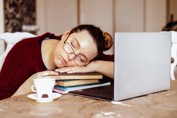 Tired young woman in glasses sleeping on books near laptop at desk in home room. Tiredness from monotonous study and rest at workplace from computer overwork. Lack of energy motivation concept