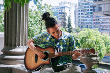 Young male, with dreadlocks hair, playing a brown acoustic guitar.