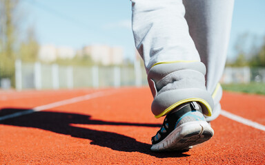 Close-up of woman athlete with fitness weights for running and weight loss training walking along track in stadium outdoors, copy space. Selective focus on leg, back view