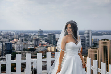 Fototapeta na wymiar Stylish gentle bride with a long veil on the roof of a tall house. A beautiful view of the city with skyscrapers and business center opens up.