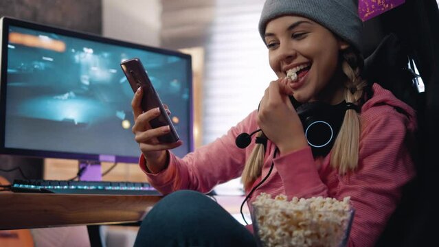 Young girl uses the phone while watching social networks and eats popcorn after playing computer games.