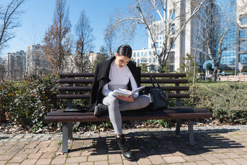 Fototapeta premium Young business woman in casual business wear reading and repeating from book preparing for job interview in the park near office building. Female sitting outdoor learning notes questions and answers.