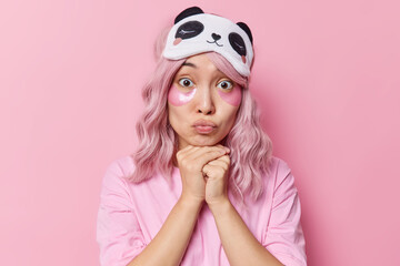 Portrait of good looking Asian woman keeps hands under chin lips folded applies beauty patches for skin treatment wears sleepmask on forehead and casual t shirt isolated over pink background