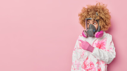 Fototapeta Shocked curly woman wears respirator and chemical suit involved in cleaning operation points away on copy space against pink background tells about protection against coronavirus and flu viruses obraz