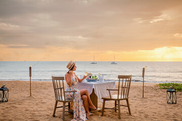 Romantic dinner on sunset. Woman sitting alone on table set with lantern for a romantic meal on beach, yachts and ocean on background. Dinner for a couple in love in luxury outdoor restaurant
