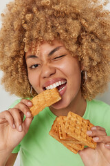 Sweet tooth concept. Close up shot of curly haired young woman bites waffles with appetite winks...
