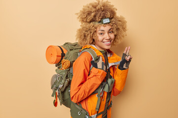 Cheerful young female camper dressed in orange jacket makes peace gesture enjoys active lifestyle carries backpack being in good mood isolated over beige background. Hiking and traveling concept