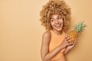 Indoor shot of happy lovely woman with curly hair holds favorite exotic fruit likes pineapples has cheerful expression looks away dressed in t shirt isolated over brown background empty space