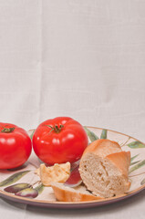 front view, medium distance of two, ripe, local, freshly picked beef steak tomatoes with a piece of baguette, hunk of cheese and a silver butter knife, on a round ceramic plate