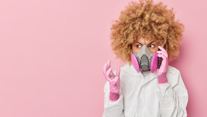 Angry young woman wears gas mask and bio hazard suit makes telephone call gestures with annoyance...