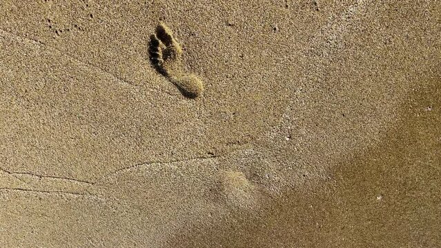 A wave of seawater washes away part of the human footprint left on the yellow sand of a sandy beach. Close-up and top view.
