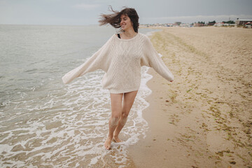 Carefree beautiful woman with windy hair running on sandy beach at cold sea waves, having fun....