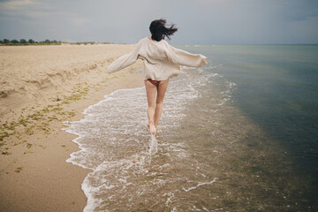 Carefree beautiful woman in knitted sweater and with windy hair running on sandy beach at cold sea,...