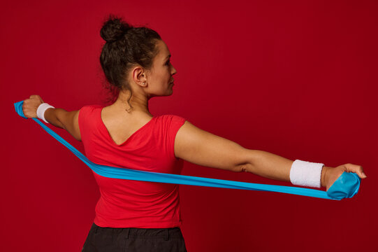 Close-up Of Beautiful Hispanic Fit Sportswoman Working Out With Elastic Rubber Strap Against Red Colored Background With Copy Ad Space