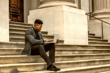 African American Businessman traveling, working in New York, wearing fashionable jacket, necktie, sitting on stairs outside office building, reading, working on laptop computer. Color filtered effect