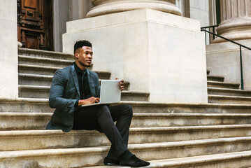 African American Businessman traveling, working in New York, wearing fashionable jacket, necktie, sitting on stairs outside office building, working on laptop computer.  filtered effect..