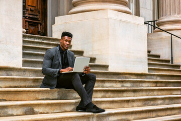 African American Businessman traveling, working in New York, wearing fashionable jacket, necktie, sitting on stairs outside office building, working on laptop computer. filtered effect..
