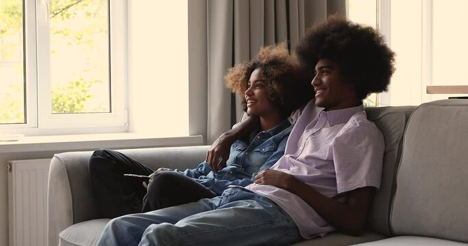 Relaxed millennial African family couple cuddle on sofa at living room hold remote controller switch on TV set enjoy funny movie comedy show. Happy young spouses spend leisure time watching television