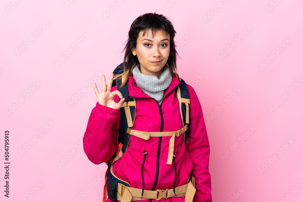 Wall mural Young mountaineer girl with a big backpack over isolated pink background showing an ok sign with fingers