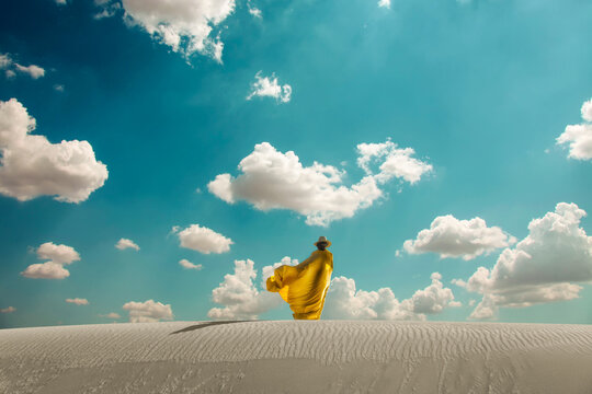 Woman In Hat And Arabic Yellow Clothes On Dune In Desert.