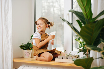 Cheerful little girl sitting on her legs on a window sill, holding spray bottle