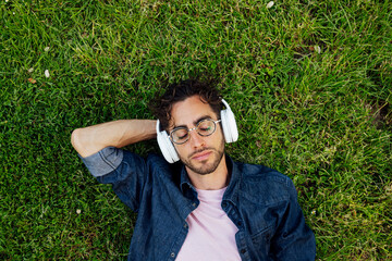 Outdoor portrait of a handsome young hipster man lying in the garden with headphones.