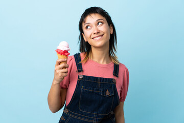 Young Uruguayan girl holding a cornet ice cream over isolated blue background looking up while smiling