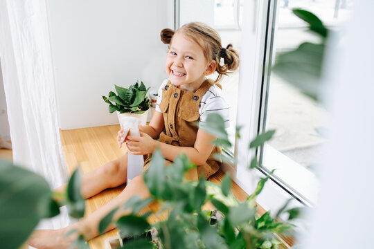 Smiling with teeth little girl sitting on a window sill, holding spray bottle