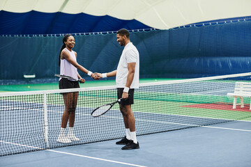 Full length shot of two tennis players shaking hands across net during match at indoor court, copy...