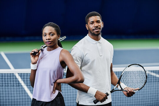 Waist up portrait of African American tennis players man and woman looking at camera while posing confidently with rackets at indoor court