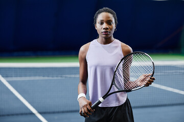 Waist up portrait of African American young woman holding tennis racket and looking at camera while posing confidently at indoor court, copy space