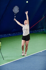 Fototapeta na wymiar Vertical portrait of young Caucasian woman playing tennis at indoor court and throwing ball in air