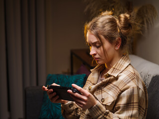 A pretty blonde girl is playing a video game on a portable game console in the room. Shooting in...