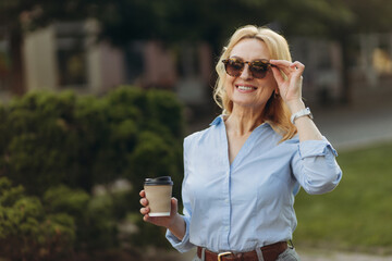 Elderly smiling happy woman 50s wearing shirt hold craft paper brown cup coffee. senior business professional walking outside
