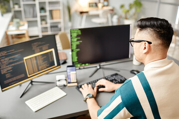 High angle view at Asian IT developer typing on keyboard with programming code on computer screen while working in office interior, copy space
