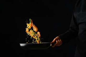 The chef cooks rice with shrimps in a frying pan on a black background. Levitation. Sea food recipes, seafood preparation. Recipe book, food blog.