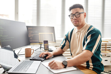 Warm toned portrait of Asian man as IT developer or QA engineer smiling at camera while sitting at...
