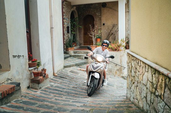 Fototapeta Cheerfully smiling man in helmet and sunglasses riding the motor scooter on the narrow Sicilian old town streets. Happy Italian vacation and transportation concept.